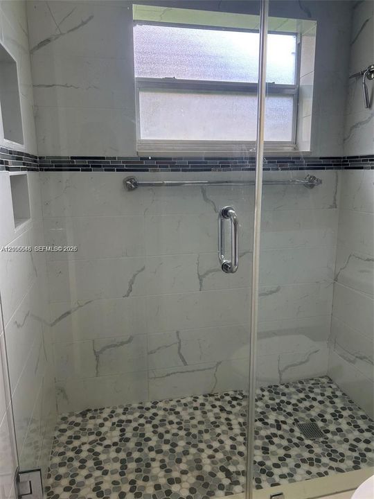 Walk-in shower featuring floor-to-ceiling stone-pattern tile, a pebble-stone floor, recessed wall niches, a decorative tile accent band, and a frameless glass door