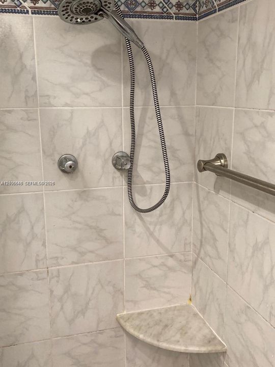 Shower enclosure featuring large-format stone-patterned wall tile, polished metal hand-held shower fixture with flexible hose, integrated corner stone shelf, and coordinating metal towel bar