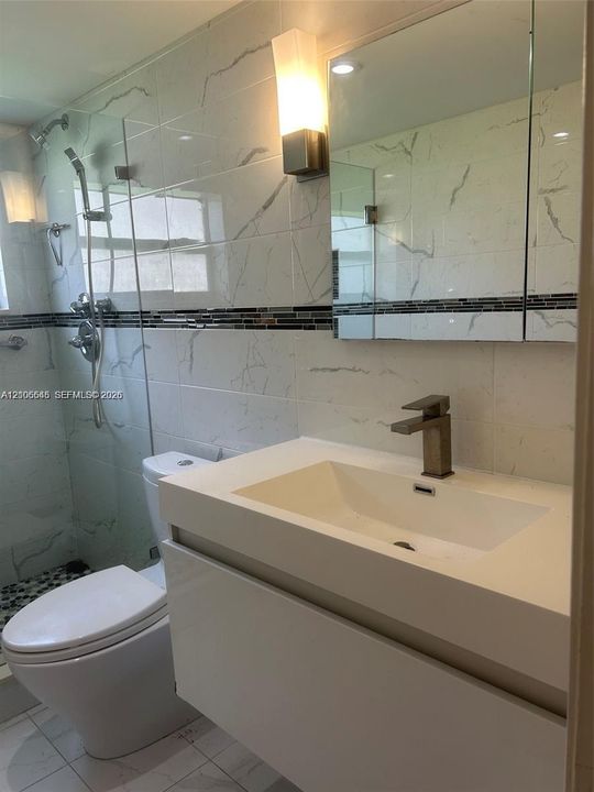 Wall-mounted vanity with a rectangular sink, stone-finish wall tile with mosaic accent border, frameless glass shower enclosure, and metallic hardware