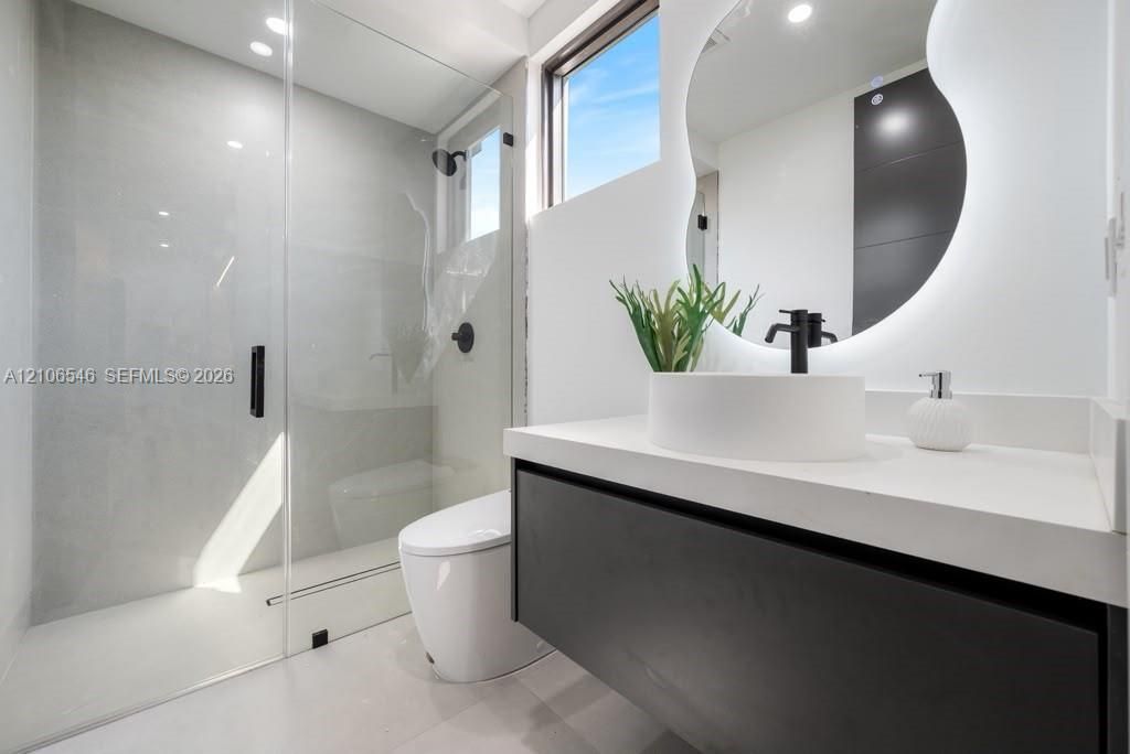 Floating dark-finish vanity with a white countertop and vessel sink, frameless glass walk-in shower with matte black fixtures, and a wall-mounted back-lit organic-shaped mirror