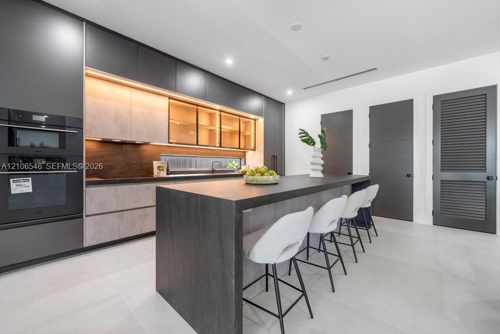 Modern kitchen featuring floor-to-ceiling dark cabinetry, a large kitchen island with a dark countertop, integrated double wall ovens, wood-finish lower cabinetry, and illuminated glass-front upper cabinets