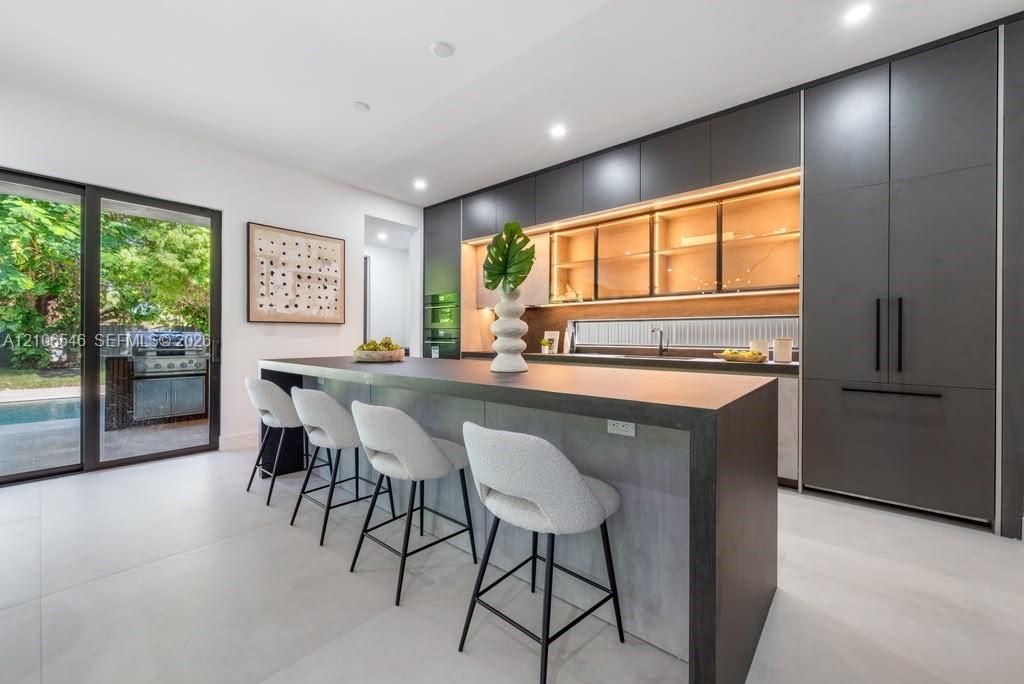 Modern kitchen featuring matte charcoal cabinetry, a large waterfall-edge island, integrated appliances, glass-front upper cabinetry with integrated lighting, and large-format porcelain tile flooring