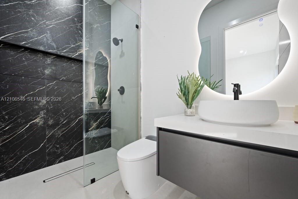 Floating vanity with white countertop and vessel sink, frameless glass shower enclosure, floor-to-ceiling dark tile shower surround, matte black hardware, and backlit organic-shaped mirror