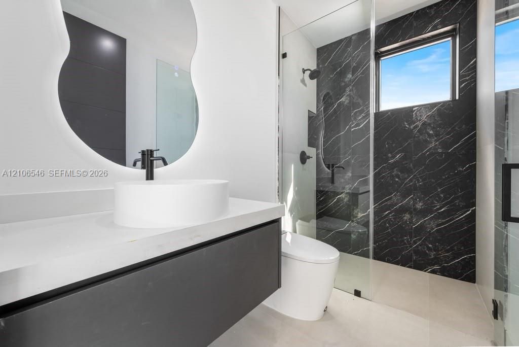 Floating vanity with a stone-composite countertop, vessel sink, backlit organic-shaped mirror, and walk-in shower featuring dark-toned tiled walls and a frameless glass enclosure