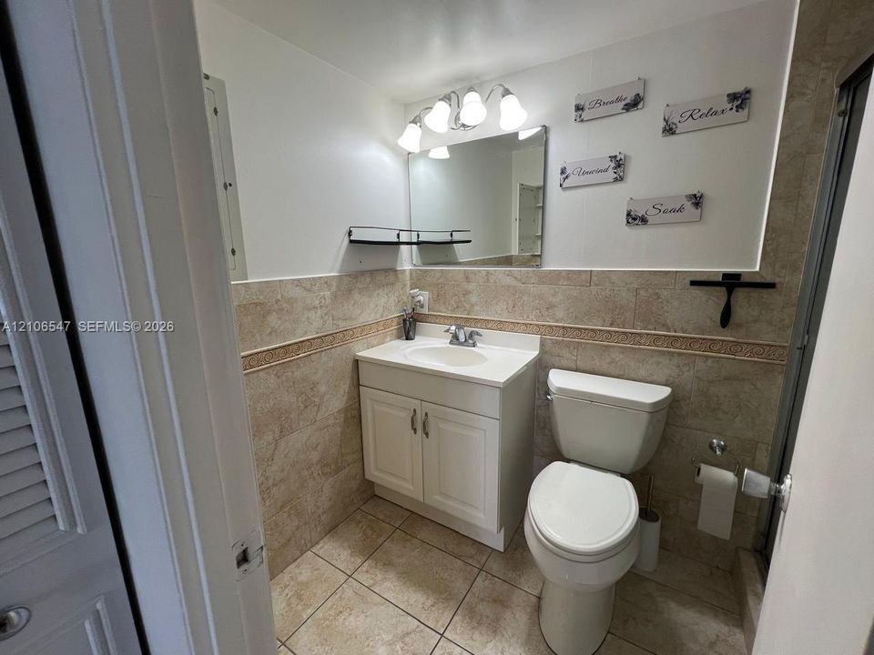 Bathroom featuring stone-finish wall and floor tile with decorative border, white cabinet vanity with integrated sink, four-light vanity fixture, and rectangular wall mirror