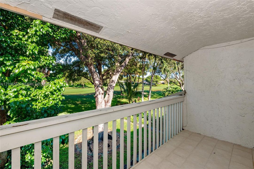 Covered balcony featuring tile flooring, textured stucco wall, and wooden balustrade railing, overlooking mature trees and golf course