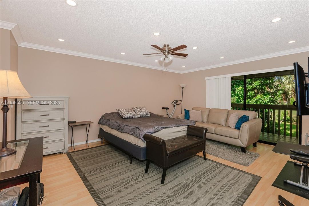 Primary bedroom. Light-toned wood-finish flooring, recessed ceiling lighting, central ceiling fan with integrated light fixture, crown molding, and sliding glass doors with access to a railed balcony