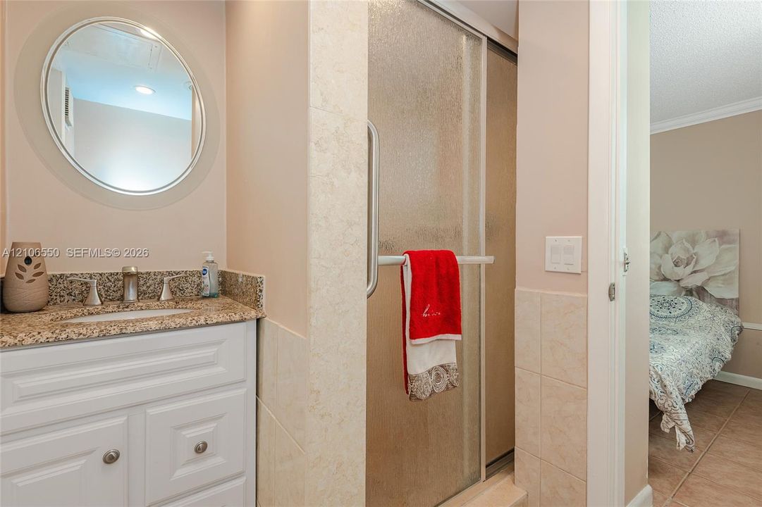 White cabinetry vanity with stone-finish countertop, undermount sink, circular wall-mounted mirror, and glass-enclosed shower with tiled surround