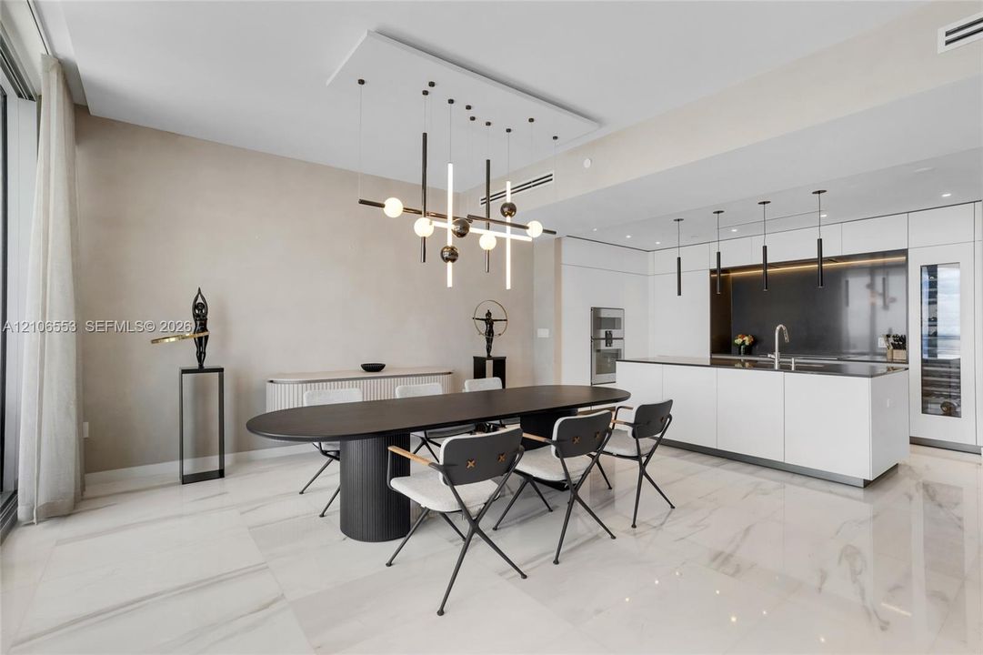 Open-concept interior featuring large-format stone flooring, a center island with integrated sink, built-in panel-ready appliances, and a geometric chandelier with linear and spherical lighting elements