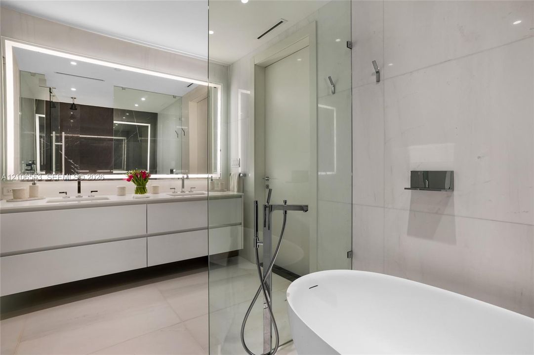 Floating vanity with dual rectangular sinks, oversized backlit mirror, floor-to-ceiling stone wall tiling, glass-enclosed shower, and freestanding soaking tub with floor-mounted faucet