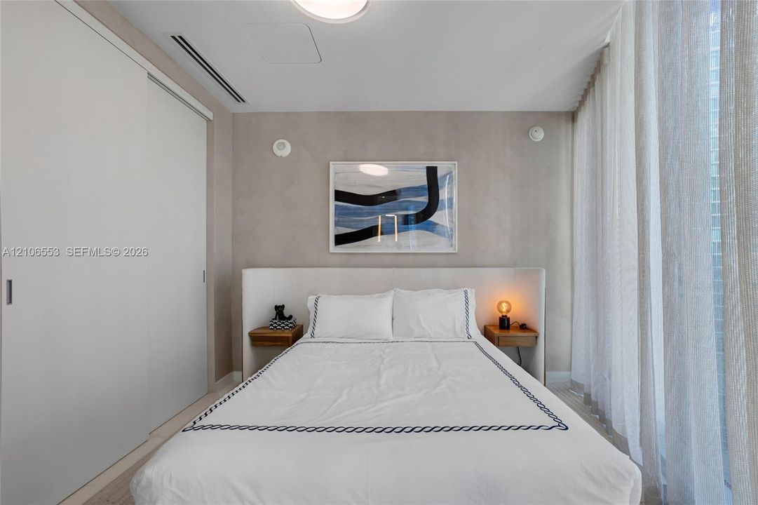 Neutral-toned room featuring a built-in upholstered headboard, dual integrated wood-finish bedside shelves, floor-to-ceiling sliding closet doors, and light-filtering floor-to-ceiling drapery