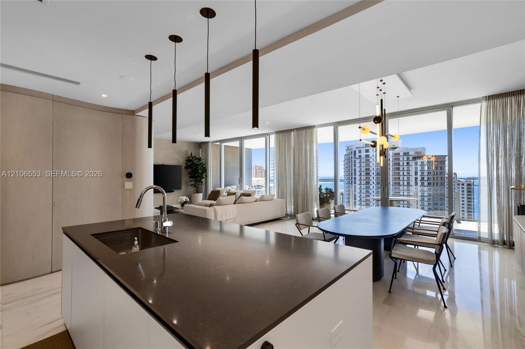 Kitchen island with a dark solid-surface countertop and undermount sink, floor-to-ceiling windows, polished stone flooring, custom cabinetry with integrated paneling, and contemporary multi-pendant lighting