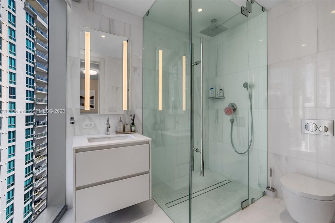 Bathroom featuring a floating vanity with a rectangular sink, wall-mounted mirror with integrated lighting, glass-enclosed shower with rainfall head and handheld sprayer, wall-mounted toilet, and floor-to-ceiling tile finishes