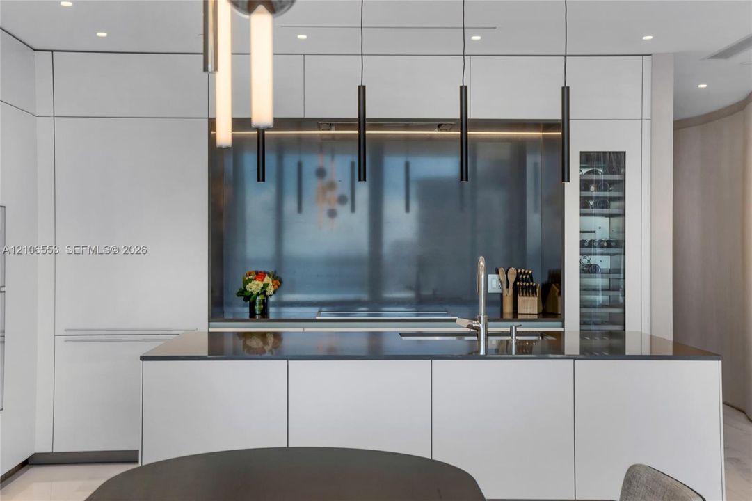 Minimalist kitchen featuring flat-panel cabinetry, a stainless steel backsplash, an integrated wine refrigerator, linear pendant lighting, and a solid-surface island with a built-in sink
