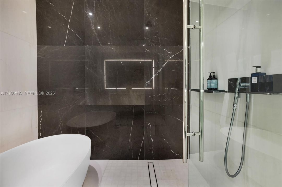 Freestanding soaking tub, floor-to-ceiling dark stone-finish wall tile, and glass-enclosed shower with polished chrome fixtures and an integrated shelf