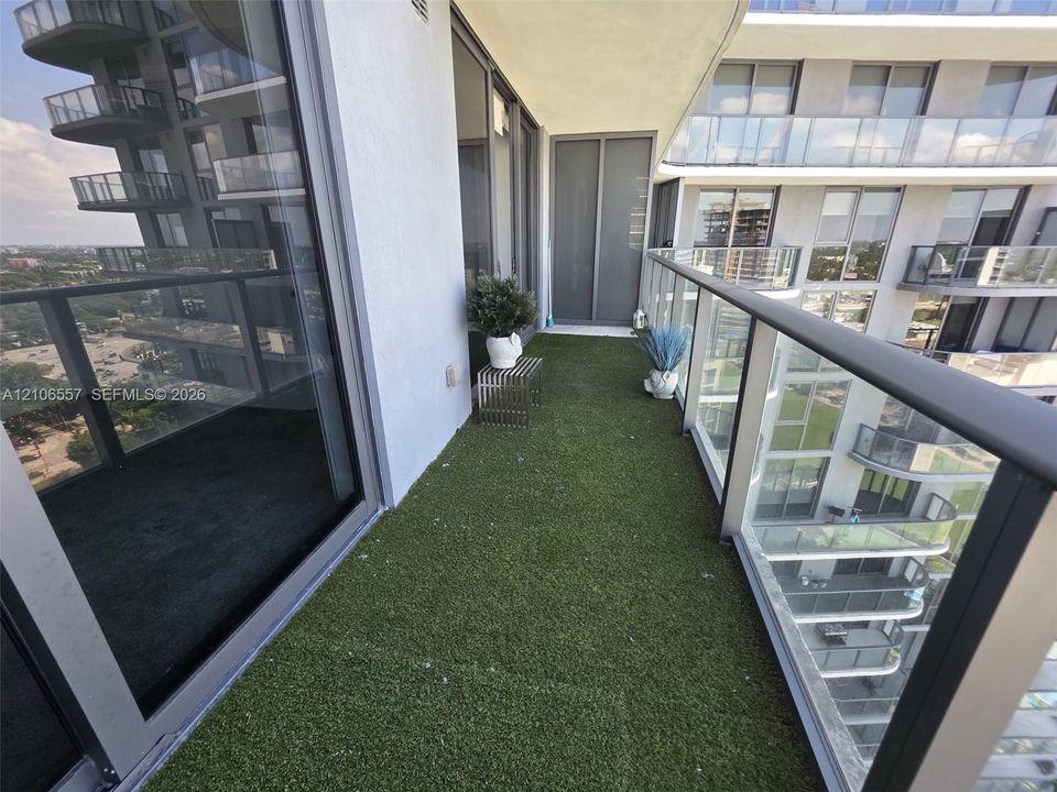 Covered balcony featuring synthetic turf flooring, floor-to-ceiling glass doors, and a glass-paneled metal railing