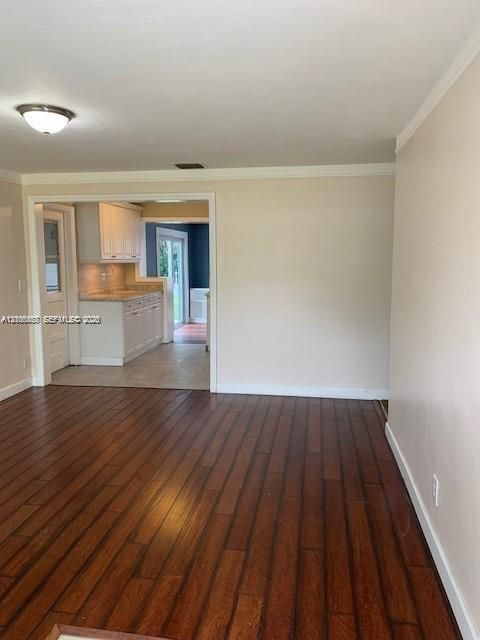 Room featuring wood-finish flooring, neutral wall paint, and crown molding, with a glimpse of a kitchen showcasing white cabinetry and stone countertops