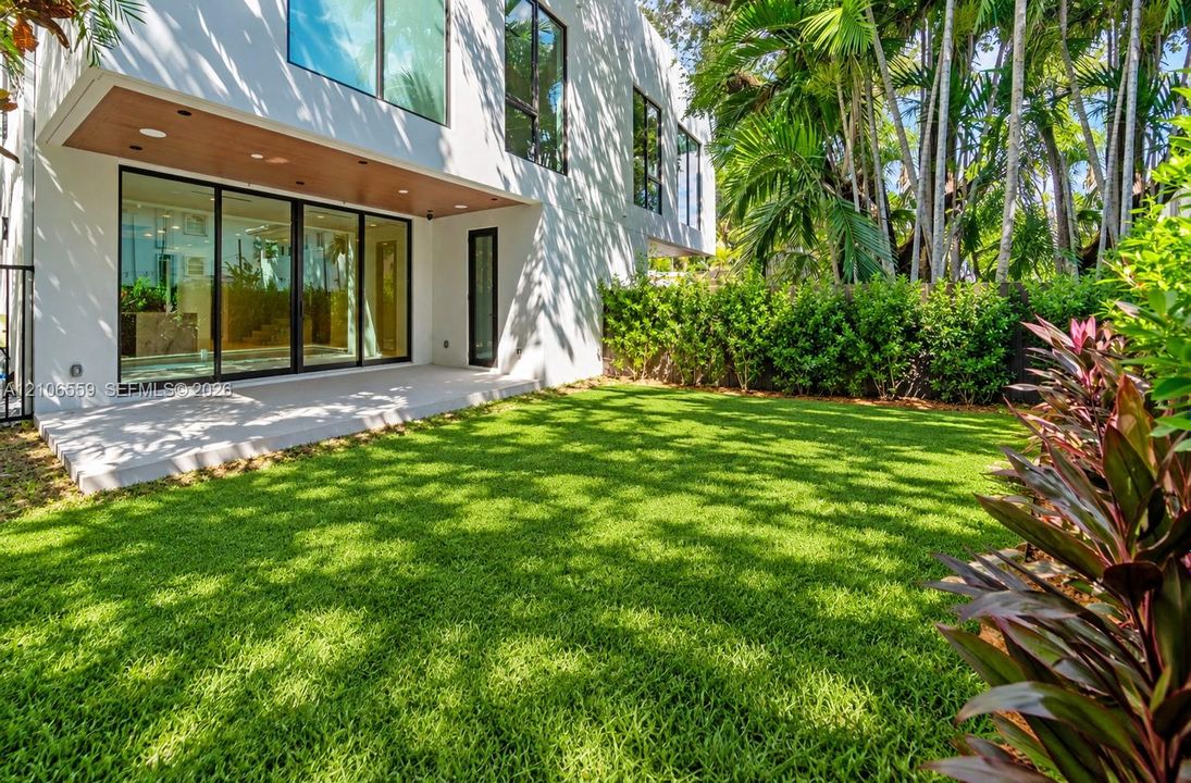 Modern architectural exterior featuring expansive glass sliding doors, wood-finish soffit, concrete patio, and fenced-in lawn with mature landscaping