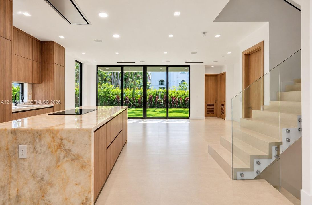 Kitchen featuring a natural stone waterfall-edge island, wood-finish cabinetry, floor-to-ceiling glass panel doors, recessed lighting, and a staircase with glass railings
