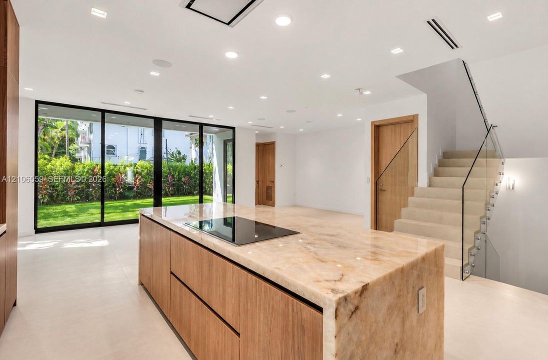 Kitchen island with natural stone countertop and integrated cooktop, floor-to-ceiling sliding glass doors, recessed lighting, and staircase featuring a glass panel railing