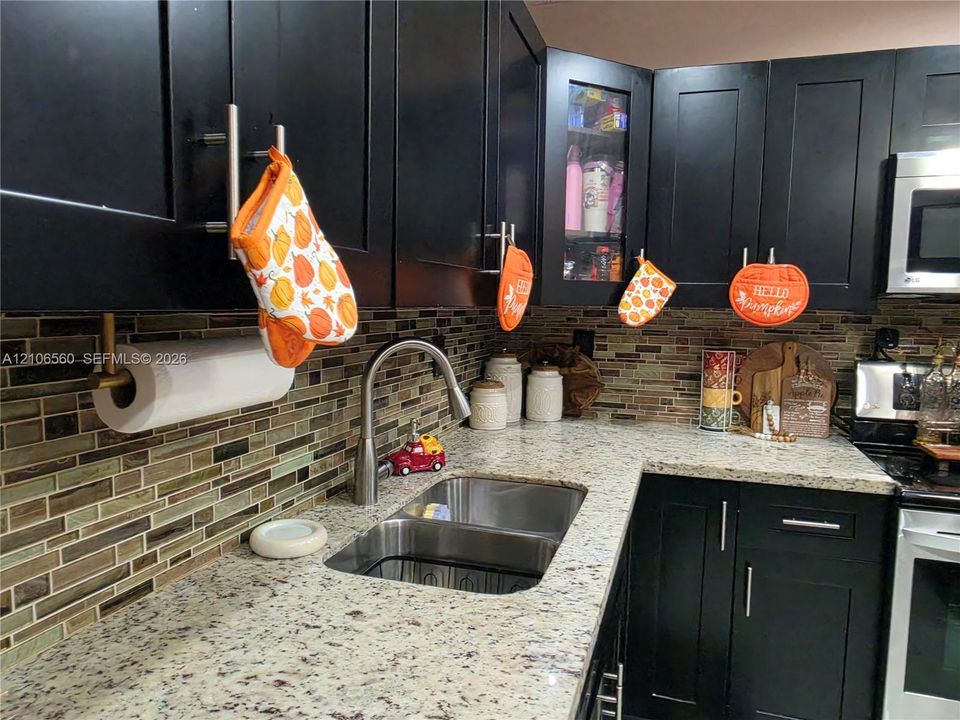 Kitchen featuring dark shaker-style cabinetry, natural stone countertops, a multi-tonal linear tile backsplash, stainless steel double-basin sink with a gooseneck faucet, and an integrated microwave