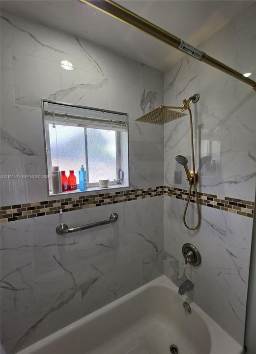 Bathtub surround featuring floor-to-ceiling stone-pattern tile, a multi-tone mosaic accent border, a gold-tone rainfall showerhead with a handheld sprayer, and a mounted metal grab bar
