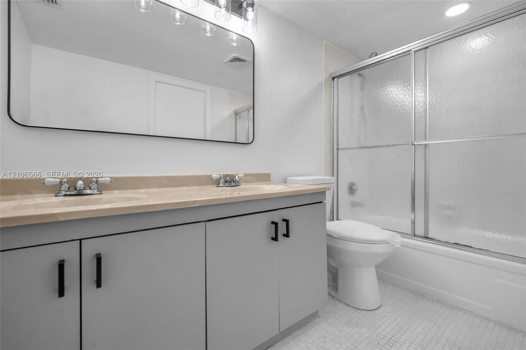Dual-sink vanity featuring neutral countertops, grey cabinetry with matte black hardware, a large rounded-corner mirror, and a shower-tub combination with sliding glass doors