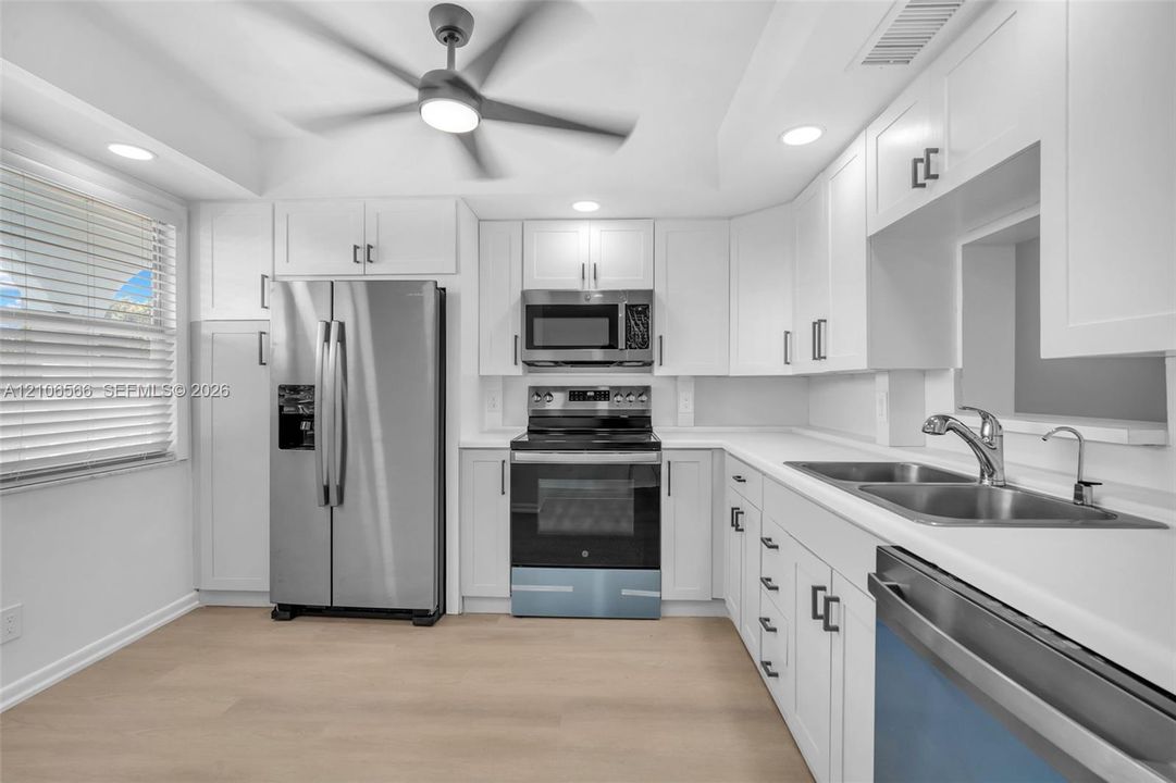 Kitchen featuring white Shaker-style cabinetry, stainless steel appliances, neutral solid-surface countertops, dual-basin stainless steel sink, and wood-finish flooring