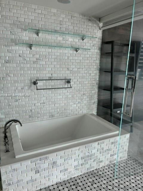 Built-in soaking tub with a tiled surround, complemented by a floor-to-ceiling accent wall in brick-pattern stone, dual glass wall-mounted shelves, and a wall-mounted metal towel rack