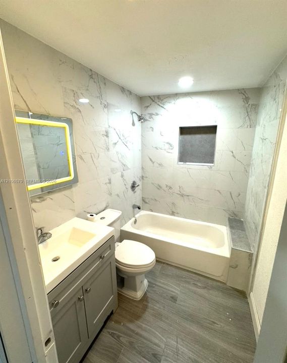 Bathroom featuring light-toned large-format wall tile, wood-finish flooring, grey cabinetry with an integrated sink, an illuminated mirror, and a tub-shower combination