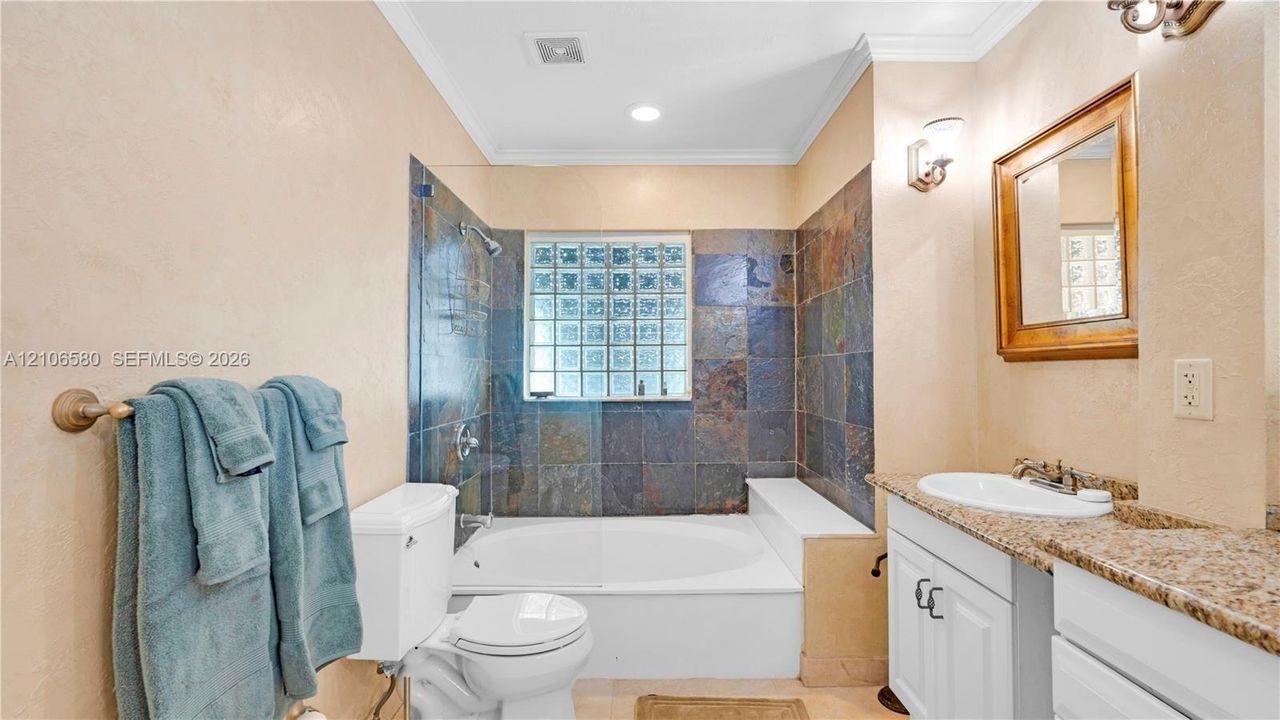 Bathroom featuring a stone-tile tub surround, glass-block window, undermount sink with a stone countertop, white shaker-style vanity cabinetry, and wall-mounted light fixtures