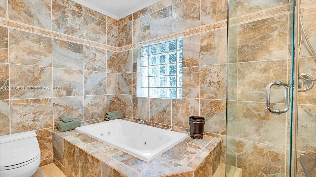 Full-wall patterned tile surround, drop-in jetted soaking tub, glass block window, and glass-enclosed shower with metallic hardware