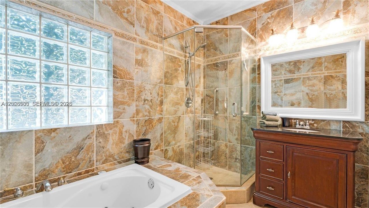Bathroom featuring full-height stone-finish wall tiling, glass block window, jetted soaking tub, corner glass-enclosed shower, and wood-finish vanity with a thick stone countertop