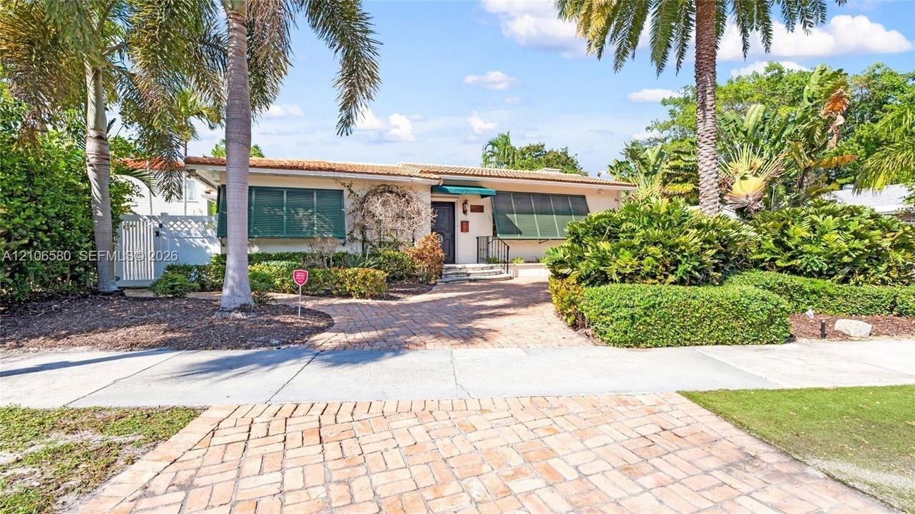 Single-story residence featuring a brick-paved driveway, clay tile roof, metal window awnings, and mature landscaping