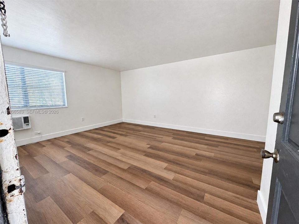 Interior space featuring wood-finish flooring, neutral wall tones, horizontal window blinds, and a wall-mounted climate control unit