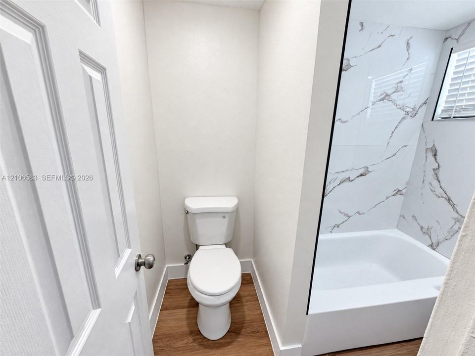 Bathroom featuring a white bathtub, tub surround with large-format veined tile, wood-finish flooring, and a standard toilet