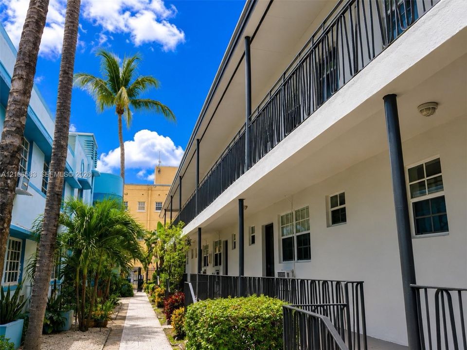 Multi-story exterior featuring a white stucco facade, metal railing, stone-paved walkway, and mature palm trees