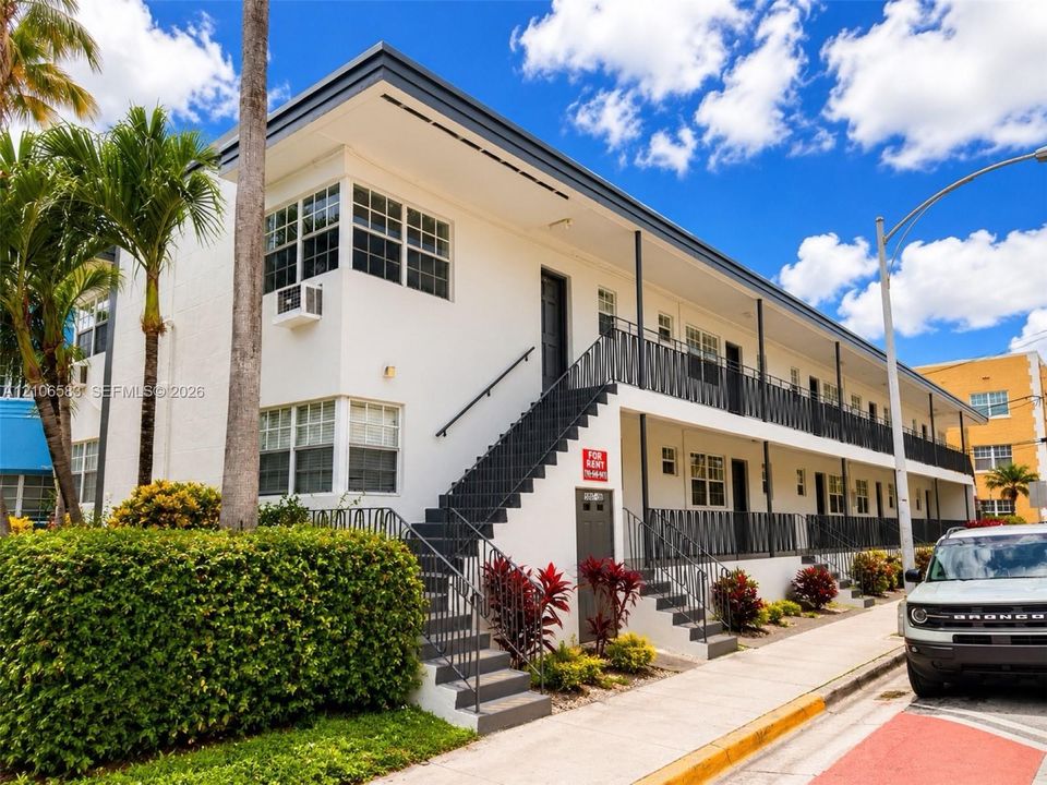 Two-story mid-century modern exterior featuring a smooth stucco finish, metal railings, exterior staircase, and mature tropical landscaping