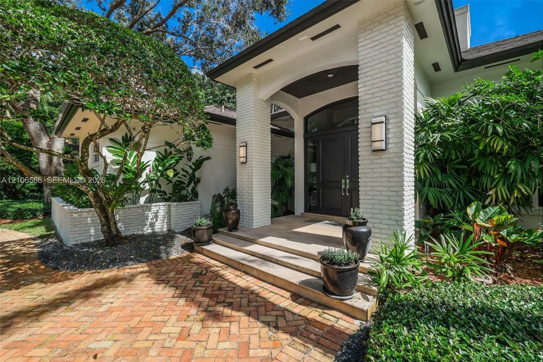 Covered entryway featuring white brick pillars, a multi-paneled dark wood front door with arched transom, tiered concrete steps, and a herringbone-patterned brick paver walkway