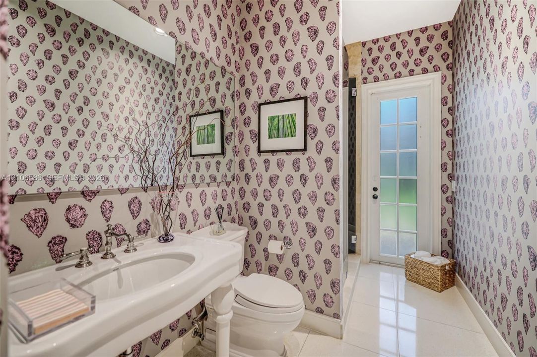 Pedestal sink with polished chrome widespread faucet, expansive wall-to-wall mirror, patterned wallpaper, and glass-paneled exterior door featuring tile flooring