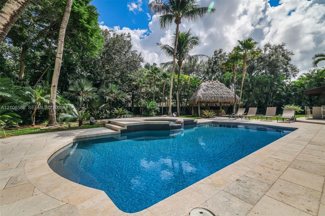 In-ground swimming pool with an integrated elevated spa, stone paver decking, and a detached thatched-roof structure surrounded by mature palms and landscaping