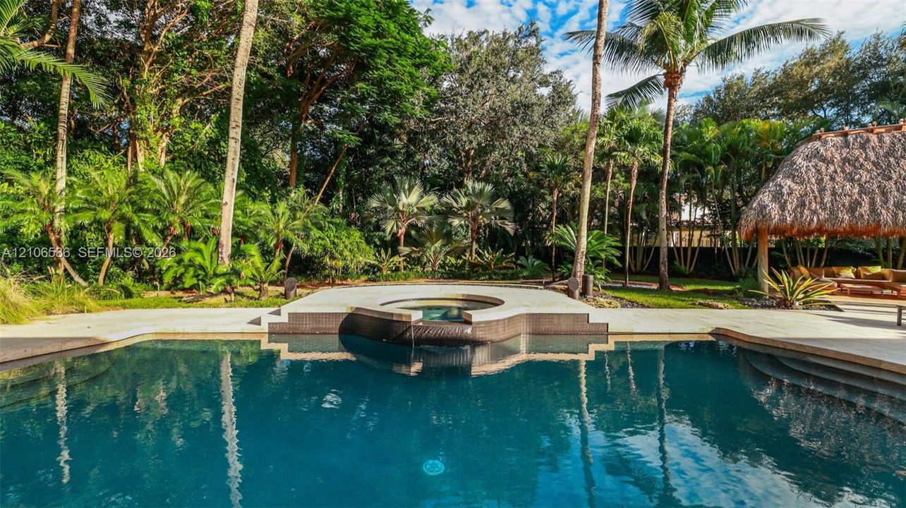 In-ground swimming pool featuring a tiered spillover spa, stone-tile surround, and a thatched-roof palapa set against a landscaped backdrop of mature palms and dense foliage