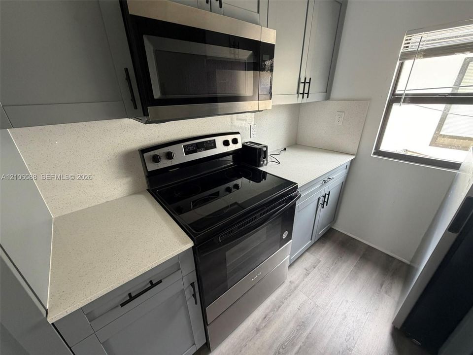 Kitchen featuring gray shaker-style cabinetry, light-toned stone-surface countertops, wood-finish flooring, and stainless steel appliances including an over-the-range microwave and electric range