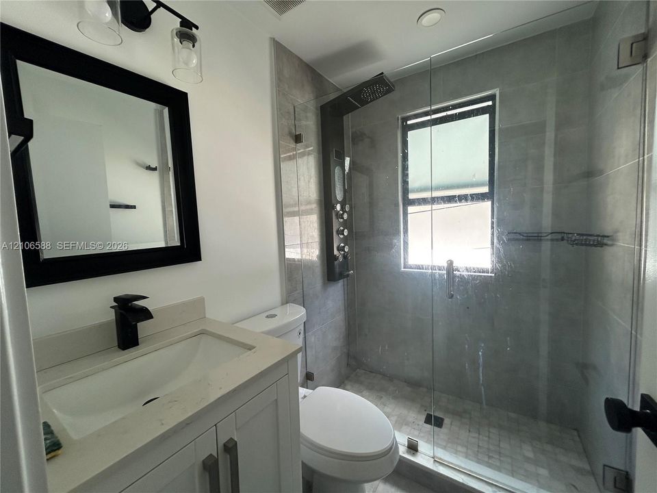 Bathroom featuring a white vanity with a composite countertop, black wall-mounted mirror, matte black faucet, glass-enclosed shower with tiled surround, and a wall-mounted shower panel system