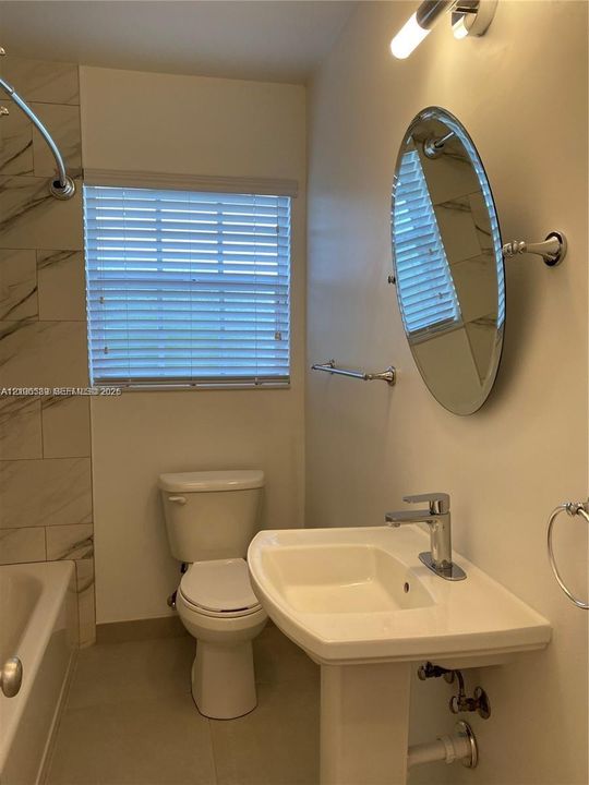 Bathroom featuring a pedestal sink, wall-mounted oval mirror, tiled tub enclosure, and neutral-toned flooring