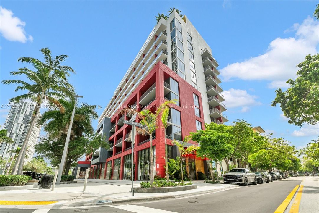 Mid-rise residential building featuring a vibrant red masonry facade, full-height glass curtain walls, private balconies, and ground-level storefronts
