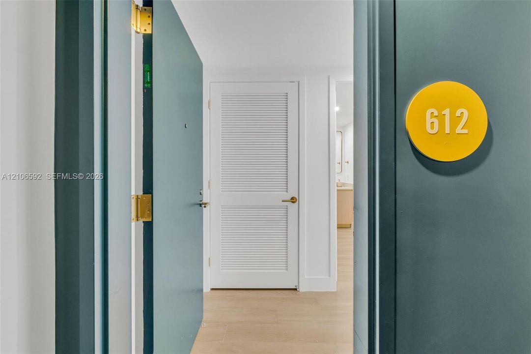 Entryway featuring a solid door with a circular yellow room identification plaque, brass hinges, white louvered door, and wood-finish flooring