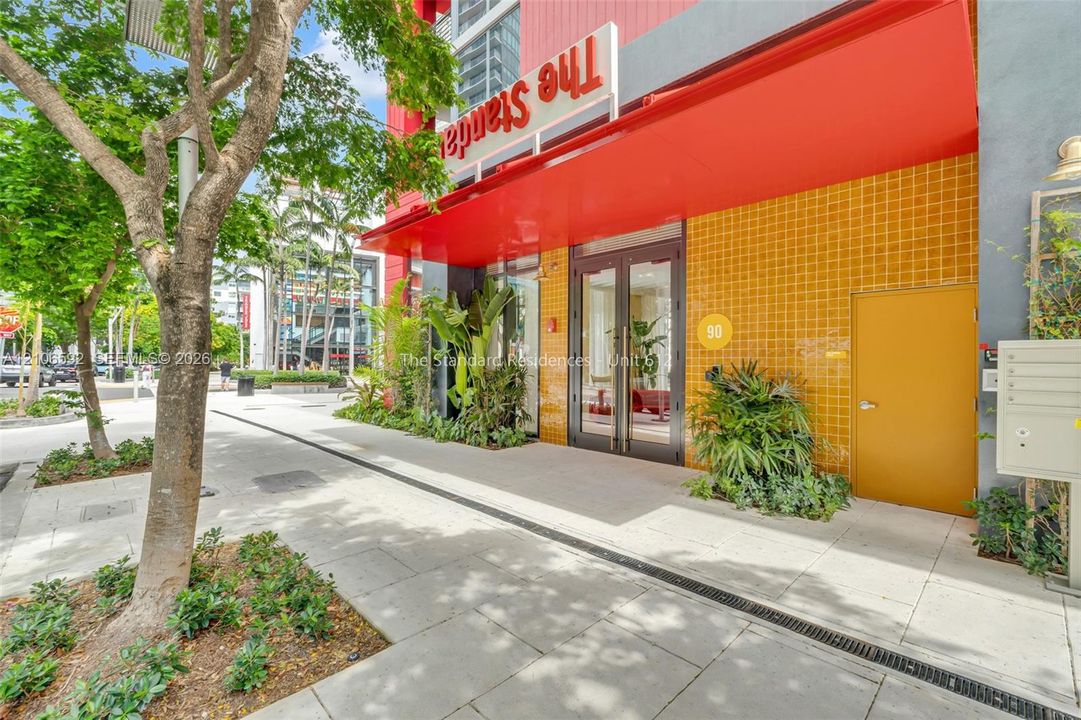 Building exterior featuring a vibrant yellow tiled facade, a prominent red metal awning, glass double-door entrance, and integrated landscape planting beds