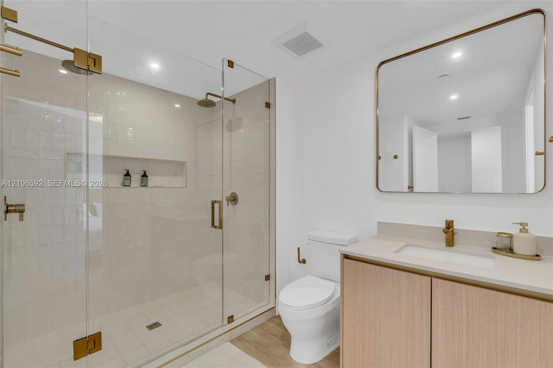 Frameless glass walk-in shower with integrated tile niche, wood-finish vanity cabinet, solid surface countertop, brass-tone hardware, and rounded-corner wall mirror