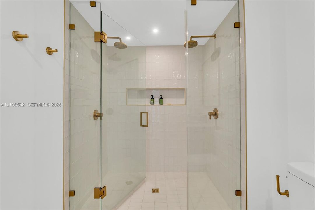 Walk-in shower featuring a frameless glass enclosure, gold-tone hardware, dual rainfall showerheads, integrated wall niche, and white subway tile wall surrounds